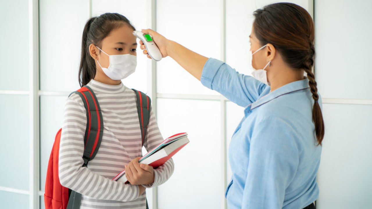 a teacher taking the temperature of a student with a thermometer before she enters the school building and schools finish reopening