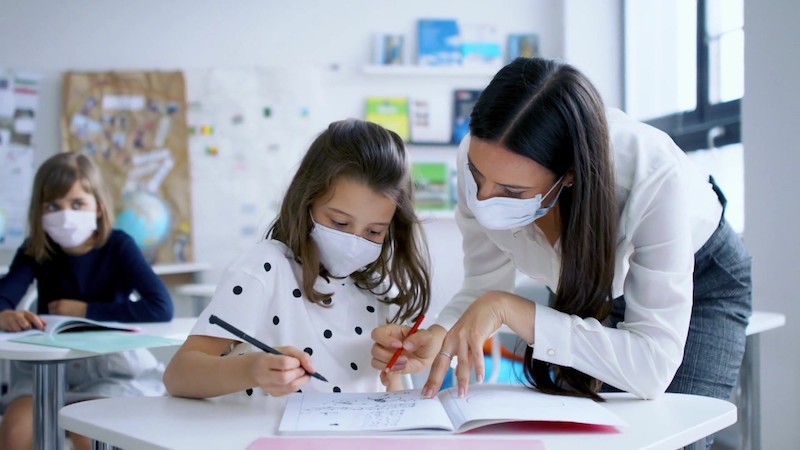 a teacher helping a student with school work as each wear masks for in-person learning 