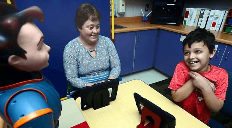 a special education student working with the milo robot and a tablet in a classroom while a therapist looks on