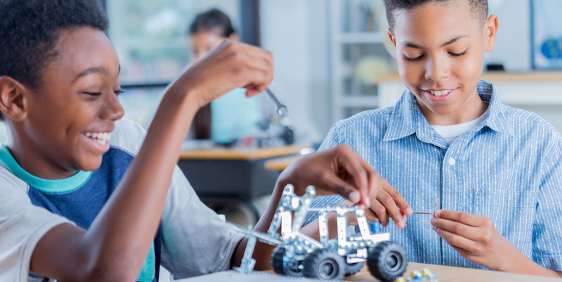 Two preteen boys sit at a desk together at school and enjoy working on a STEM robotics project together.