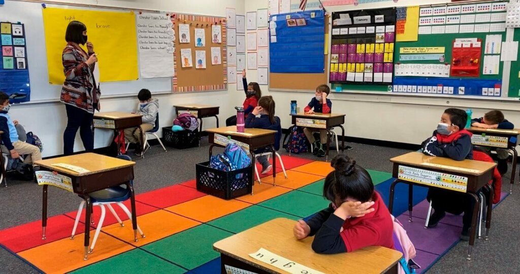 students in masks sitting in a classroom as teachers run a lesson