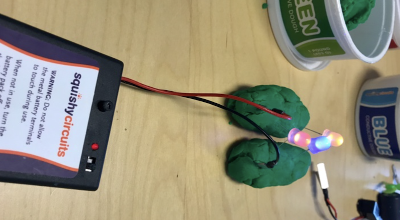 a squishy circuits project consisting of two green piles of conductive dough, batteries, wires, and illuminated LEDs
