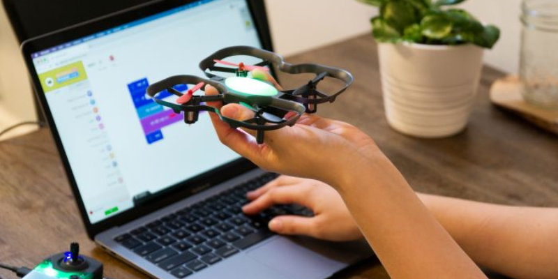 a student holding a small drone in his hand and writing a program for controlling its flight path on a laptop