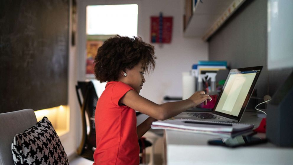 a young child sits at a bedroom desk with a laptop open for school work